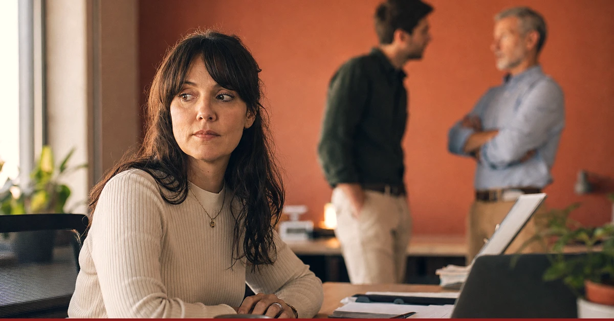 Eine neue Teamleiterin sitzt am Schreibtisch und sieht, wie ein Mitarbeiter im Hintergrund direkt mit der Geschäftsführung spricht, an ihr vorbei.